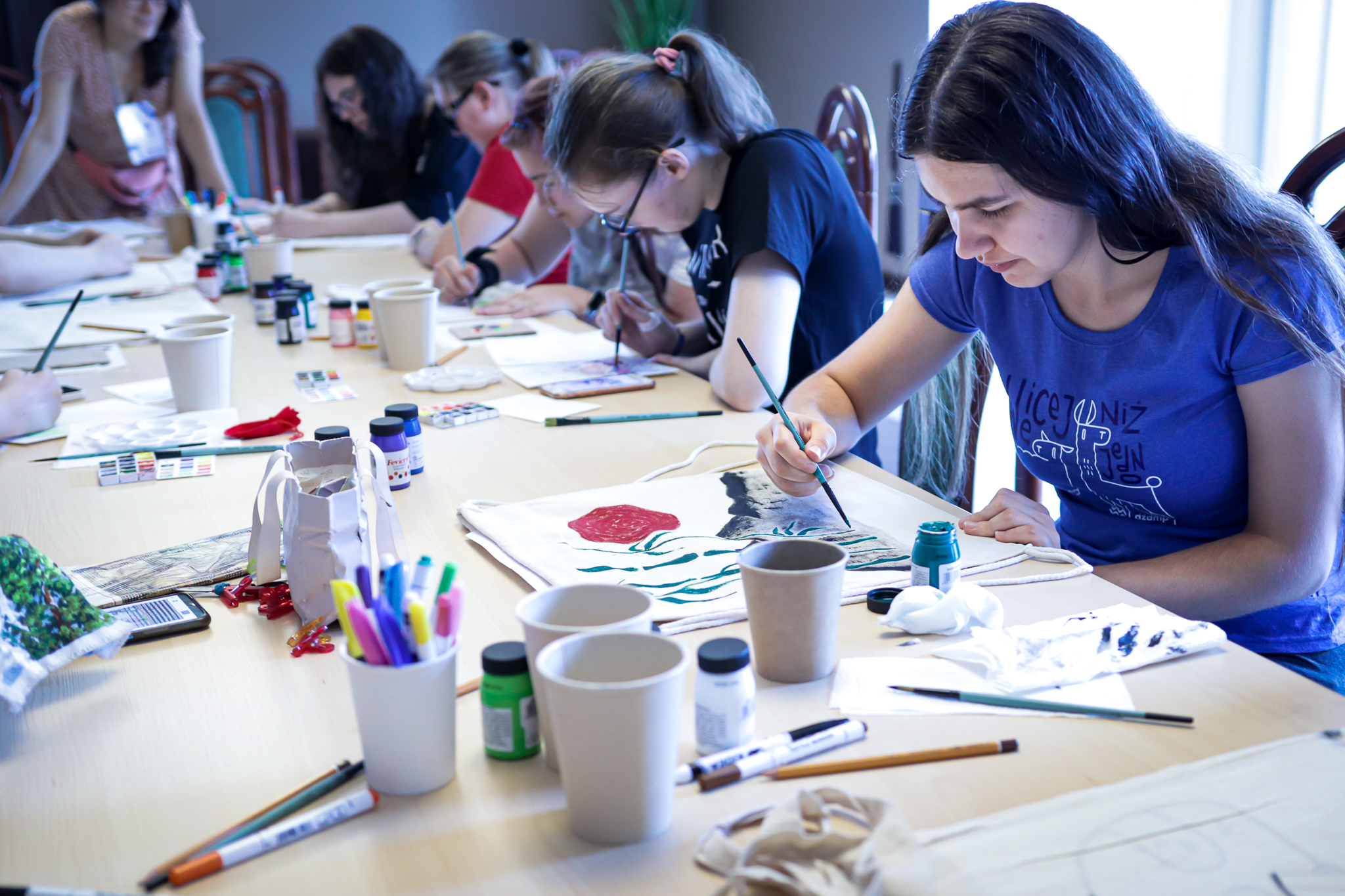 Kilka osób siedzących przy stole i malujących torby farbami.